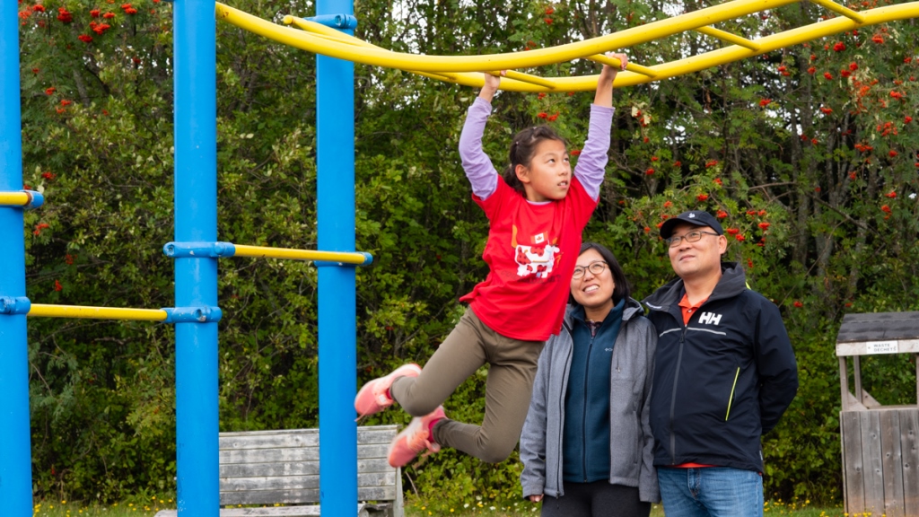 A young girl playing on monkey bars at a playground while her parents stand nearby, smiling and watching her. 