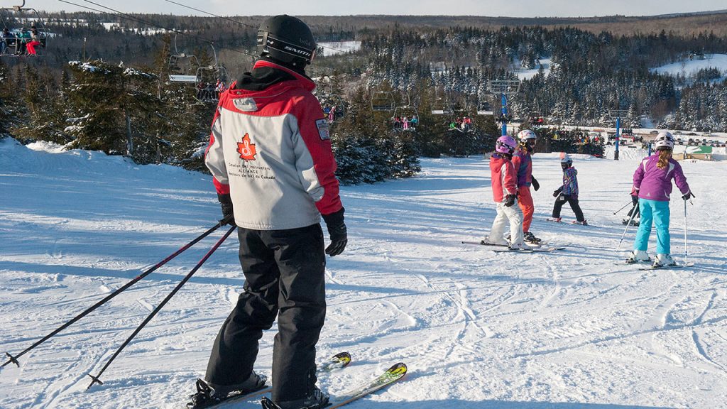 A ski instructor standing on a snowy slope, observing a group of young skiers at a ski resort.